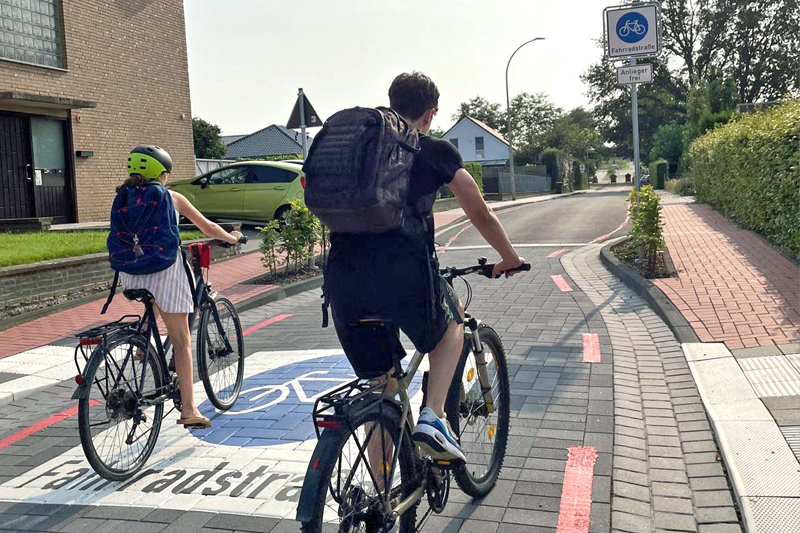 Eine Schülerin und ein Schuler fahren mit dem Rad auf einer Fahrradstraße. Im Vordergrund ist die Markierung "Fahrradstraße" auf der Fahrbahn aufgebracht, im Hintergrund ein entsprechendes Schild.