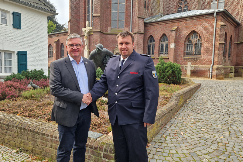 Bürgermeister Karl-Hein Wassong gibt dem stellvertrenden Wehrleiter Jens Claßen die Hand. Im Hintergrund die Kirche St. Laurentius in Elmpt. Links daneben das weiße "Haus Hansen"