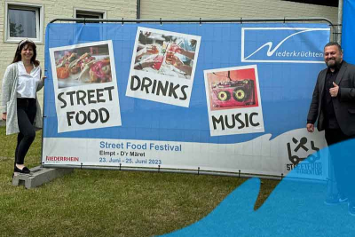 Nadine Buscher (Gemeinde Niederkrüchten) und Tuncay Ulupinar (EnMa Deutschland) stehen vor einem Banner mit der Ankündigung des ersten Street Food Festivals in Elmpt