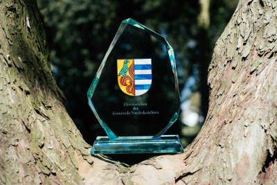 Der gläserne Pokal des Ehrenzeichens der Gemeinde Niederkrüchten steht auf einem Baumstumpf. Auf dem Pokal ist das Logo der Gemeinde Niederkrüchten aufgedruckt.