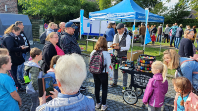 Ein Ballonkünstler steht vor einem Pavillon der Gemeinde Niederkrüchten. Um ihn herum stehen Kinder und Erwachsene, die ihm zusehen.