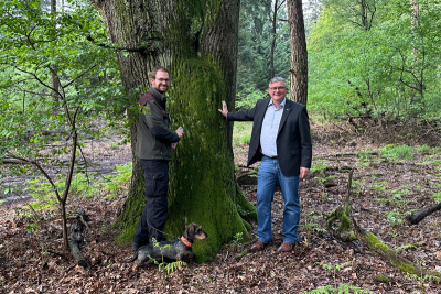 Gemeindeförster Simon Gossen, Bürgermeister Karl-Heinz Wassong und Dackel Arthur vor einem Baum im Wald