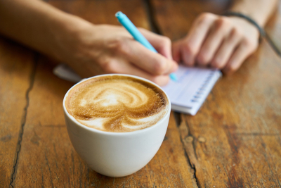 Eine Tasse Café auf einem Holztisch. Im Hintergrund wird mit einem hellblauen Stift auf einen Notizblock geschrieben.