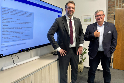 Die Bürgermeister der Gemeinden Schwalmtal und Niederkrüchten, Andreas Gisbertz und Karl-Heinz Wassong vor einem großen Bildschirm, auf dem eine Ausgabe des Chatbots zu sehen ist.