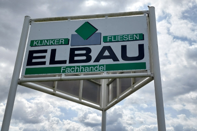 Schild mit dem Logo der Elbau Jansen GmbH & Co. KG. Im Hintergrund leicht bewölkter Himmel.